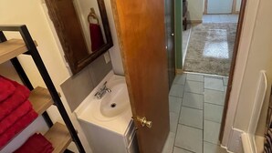 Une salle de bain avec un lavabo blanc, un miroir et des serviettes rouges. Une porte en bois donne sur un couloir dont le sol est recouvert d’une moquette.