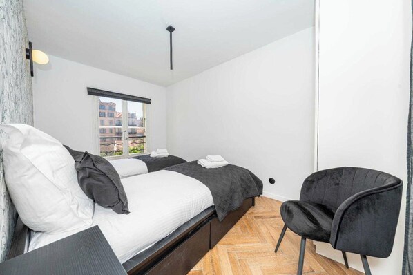Une chambre avec deux lits, une chaise noire et une fenêtre donnant sur des immeubles.