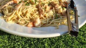 Eine Pasta-Kombination mit Garnelen und Hähnchen auf einem weißen Teller, platziert auf einer grasbewachsenen Fläche mit einem gelben Gebäude im Hintergrund.