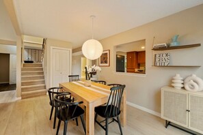 A dining area with a wooden table and chairs, a hanging light fixture, and a kitchen visible through an open door. - Chic 3BR Home | Foosball • Great Location@Bellevue (Bellevue)