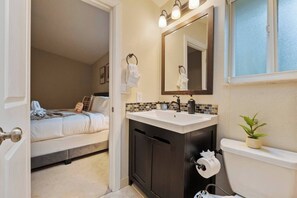 Ein Badezimmer mit Waschbecken, Spiegel und Toilette. Im angrenzenden Raum befindet sich ein Bett.