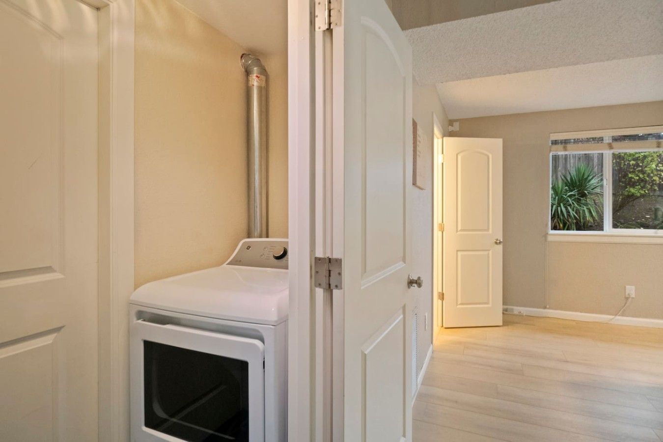 A laundry room with a washer and dryer, a door leading to another room, and a window with a view of greenery.