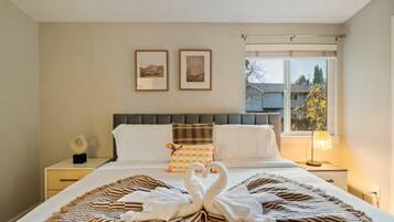 A hotel room with a bed, two framed pictures on the wall, a window with a view, and a bedside lamp.