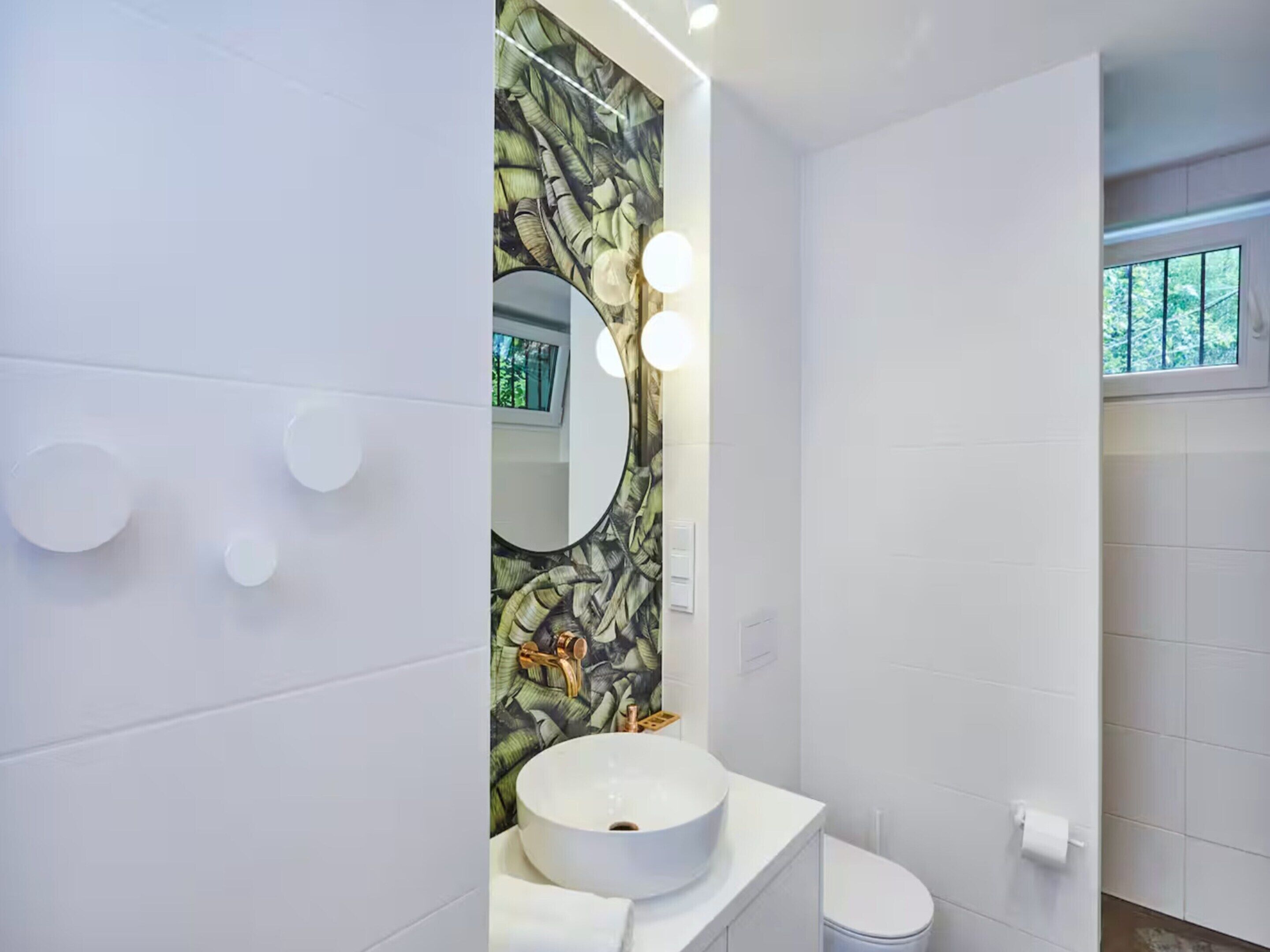 A modern bathroom with a round sink, a mirror with lights, and a window with a view of trees.