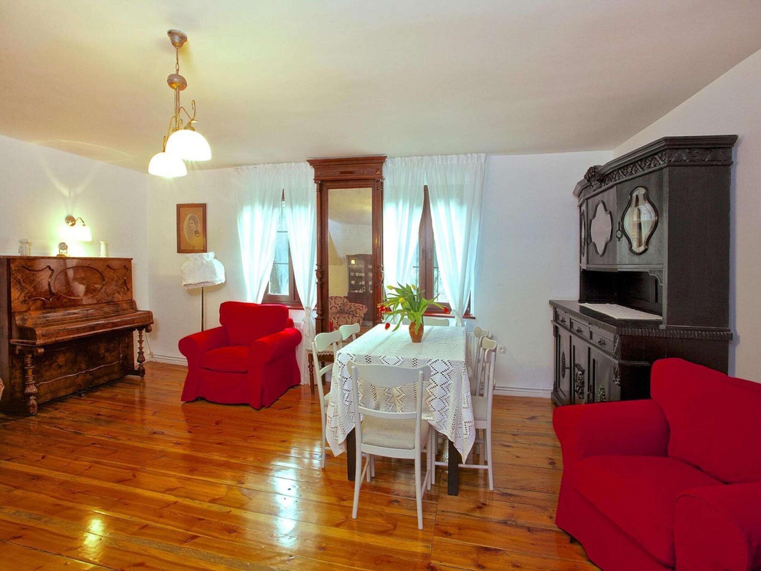 Un soggiorno con pavimento in legno, una poltrona rossa, un tavolo da pranzo con tovaglia bianca, un divano rosso, un armadio in legno e un pianoforte.