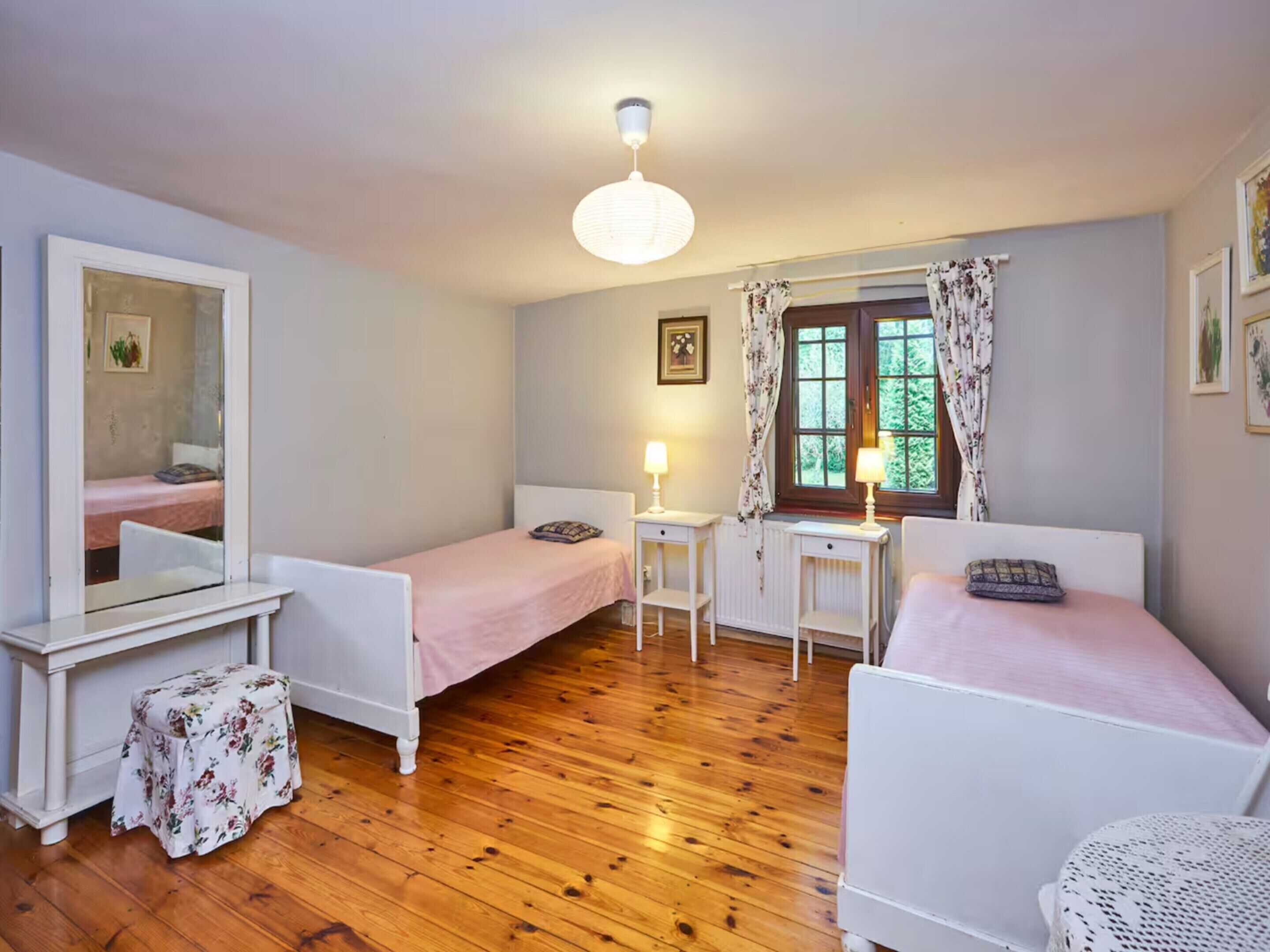 A small, well-lit room with two single beds, a desk, a chair, a mirror, and a window with curtains.