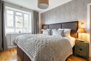 Une chambre à coucher avec un grand lit, une table de chevet, une lampe et une fenêtre avec des rideaux.
