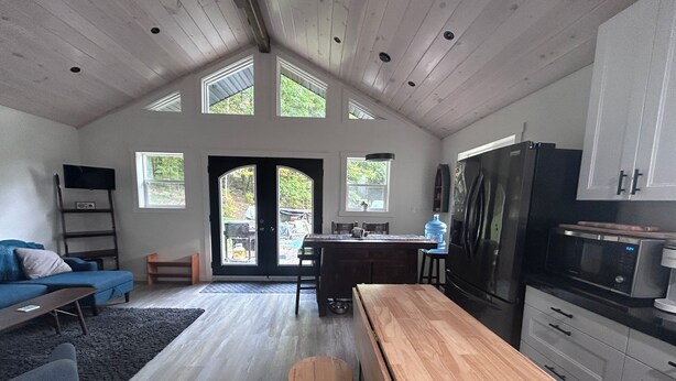 A spacious living area with a wooden dining table, a kitchen with modern appliances, and a large window overlooking a garden. - Charming Muskoka Wilderness Cottage (Windermere)