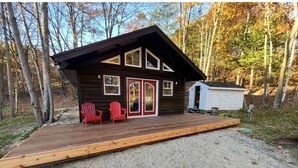 A cabin with a wooden deck, a red chair, and a white shed in a wooded area. - Charming Muskoka Wilderness Cottage (Windermere)
