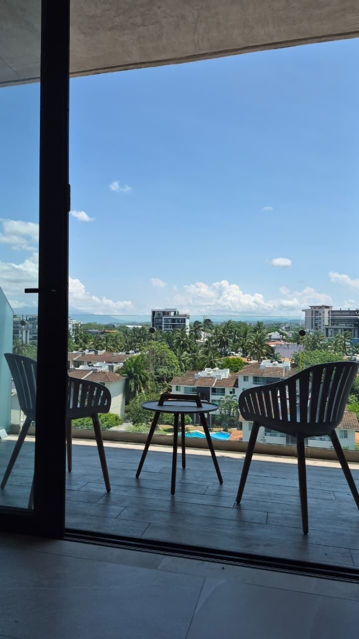 Un balcón con dos sillas y una mesa, con vista a una zona residencial con casas y cielo despejado.