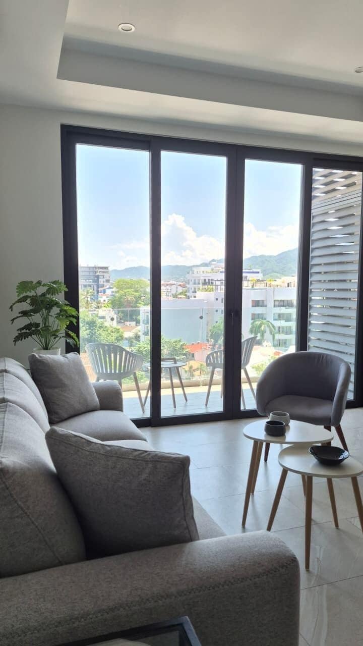 Een moderne woonkamer met een bank, een stoel en een kleine tafel. Er is een groot raam met uitzicht op gebouwen en groen.