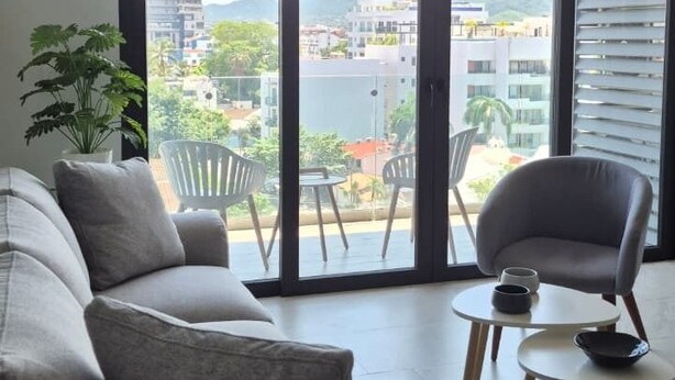 A modern living room with a sofa, a chair, and a small table. There is a large window with a view of buildings and greenery. - Relax, Unwind and Experience Vallarta (Puerto Vallarta)