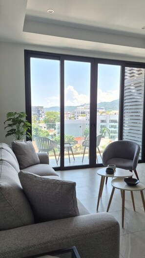 A modern living room with a sofa, a chair, and a small table. There is a large window with a view of buildings and greenery. - Relax, Unwind and Experience Vallarta (Puerto Vallarta)