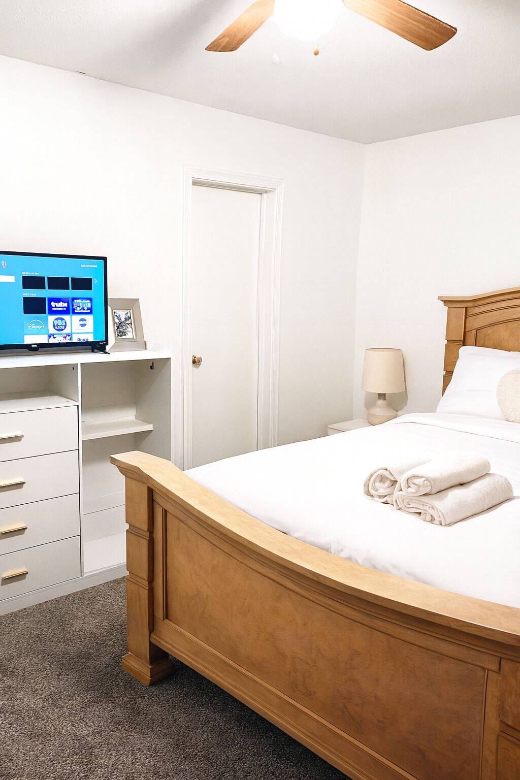 Un dormitorio con cama de madera, paredes blancas, televisor sobre un mueble, una puerta y una mesita de noche con lámpara.