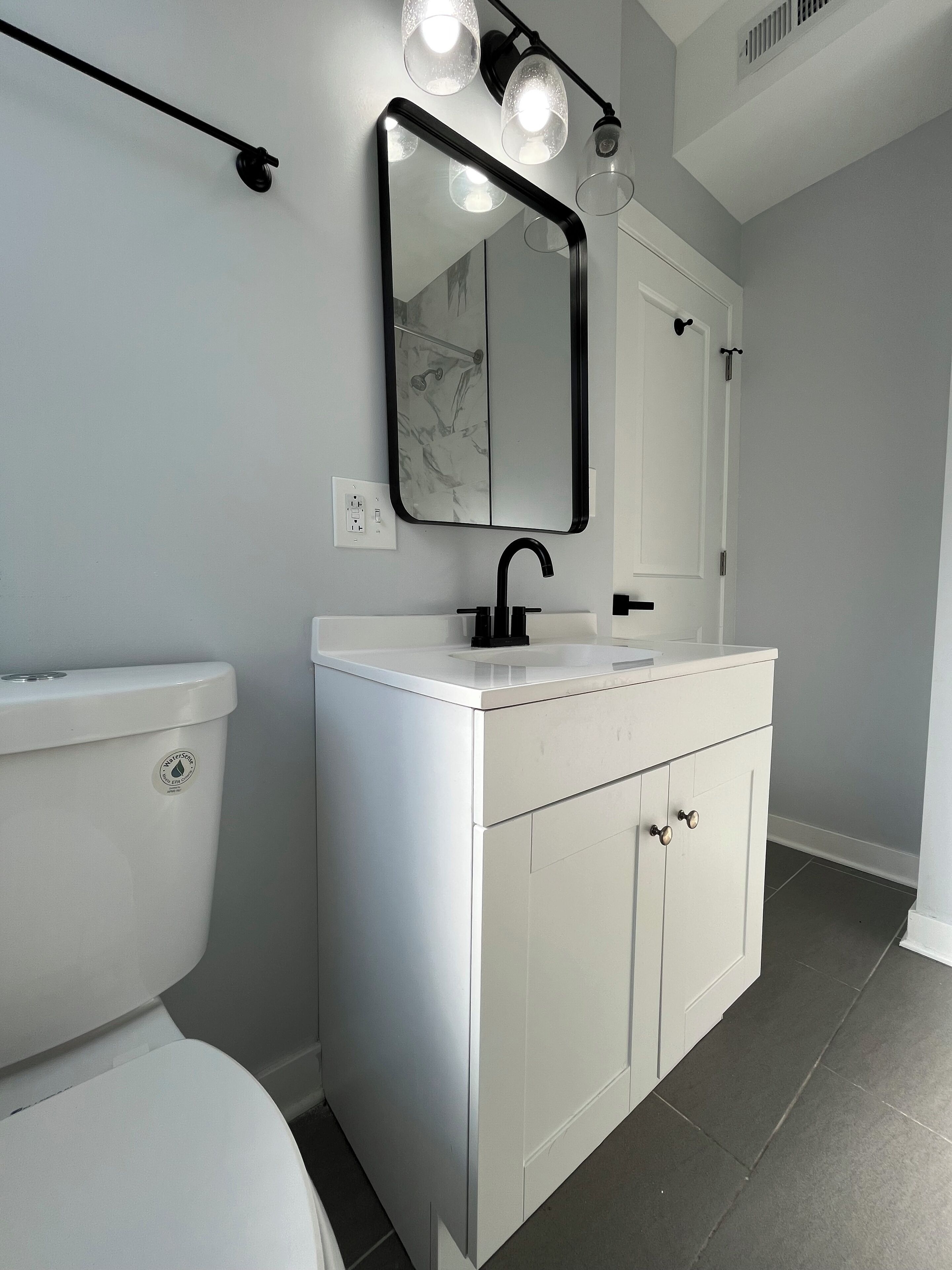 A modern bathroom with a white toilet, a white vanity with a black faucet, and a mirror with three lights.