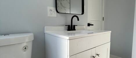 A modern bathroom with a white toilet, a white vanity with a black faucet, and a mirror with three lights.