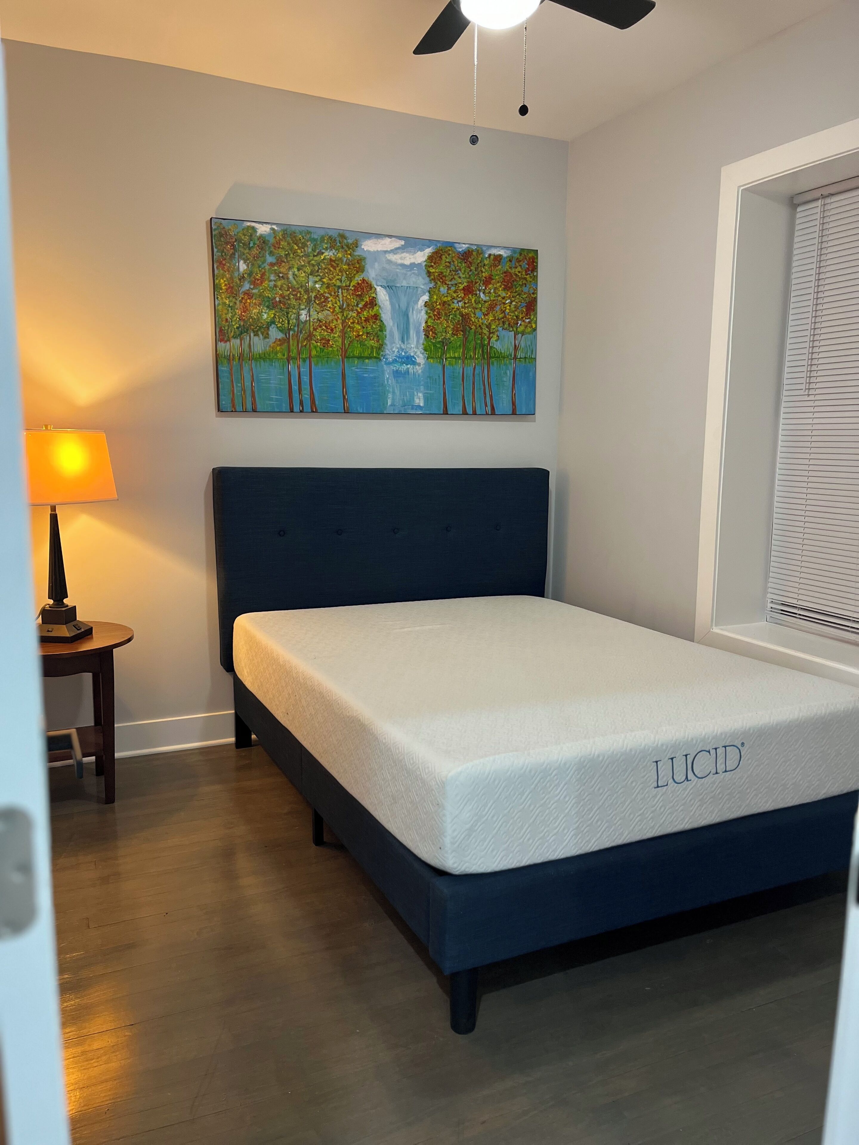 Une chambre à coucher avec un lit, un tableau représentant une cascade, une lampe et une fenêtre avec des stores.