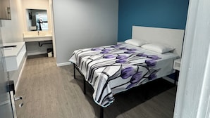 A bedroom with a bed, a bedside table, a nightstand, a mirror, and a bathroom with a sink and mirror. - Angel Motel near Anaheim  (Orange)