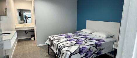 A bedroom with a bed, a bedside table, a nightstand, a mirror, and a bathroom with a sink and mirror.