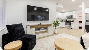 Un salon moderne avec un téléviseur à écran plat, un canapé noir, des tables d’appoint en bois et une cuisine en arrière-plan.