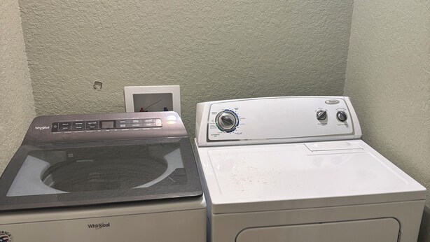 Een ruimtelijk wasgedeelte met een voorlader wasmachine en droger, een klein wit kastje en een structuurwand.