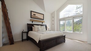 Ein Schlafzimmer mit großem Fenster, einer Holzleiter, einem Bett mit Quilzdecke und einem gerahmten Bild an der Wand.