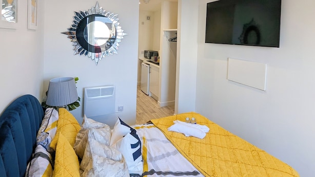 Hotelkamer met een bed, een aan de muur bevestigde televisie, een spiegel en een kleine kitchenette op de achtergrond.
