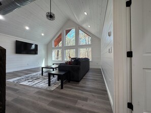 A spacious loft with a high ceiling, a large window with a view of trees, a black leather sofa, and a flat-screen TV mounted on the wall. - Gorgeous New Skyfall Cabin in Broken Bow Sleeps 10 in Style with Huge Deck! (Broken Bow)