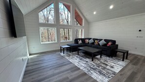 Een loft-achtige kamer met schuin plafond, grote ramen, een hoekbank, een salontafel en een kamerbreed tapijt met een patroon.