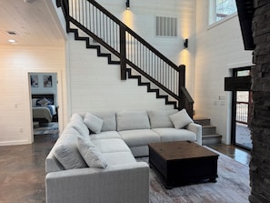 A spacious living room with a large L-shaped gray sectional sofa, a dark wooden coffee table, and a black staircase with a railing. - Gorgeous New Skyfall Cabin in Broken Bow Sleeps 10 in Style with Huge Deck! (Broken Bow)
