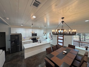 A modern kitchen with white cabinets, a stainless steel refrigerator, a microwave, and a large island with a sink. There is a dining area with a table and chairs, and a chandelier above. - Gorgeous New Skyfall Cabin in Broken Bow Sleeps 10 in Style with Huge Deck! (Broken Bow)