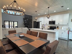A modern kitchen with white cabinets, a central island, and a dining area with a dark table and brown chairs. - Gorgeous New Skyfall Cabin in Broken Bow Sleeps 10 in Style with Huge Deck! (Broken Bow)