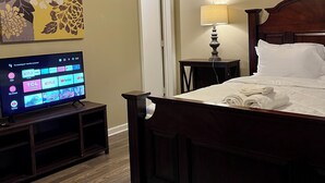 A hotel room with a bed, a TV on a stand, a ceiling fan, a lamp, and a floral artwork on the wall.