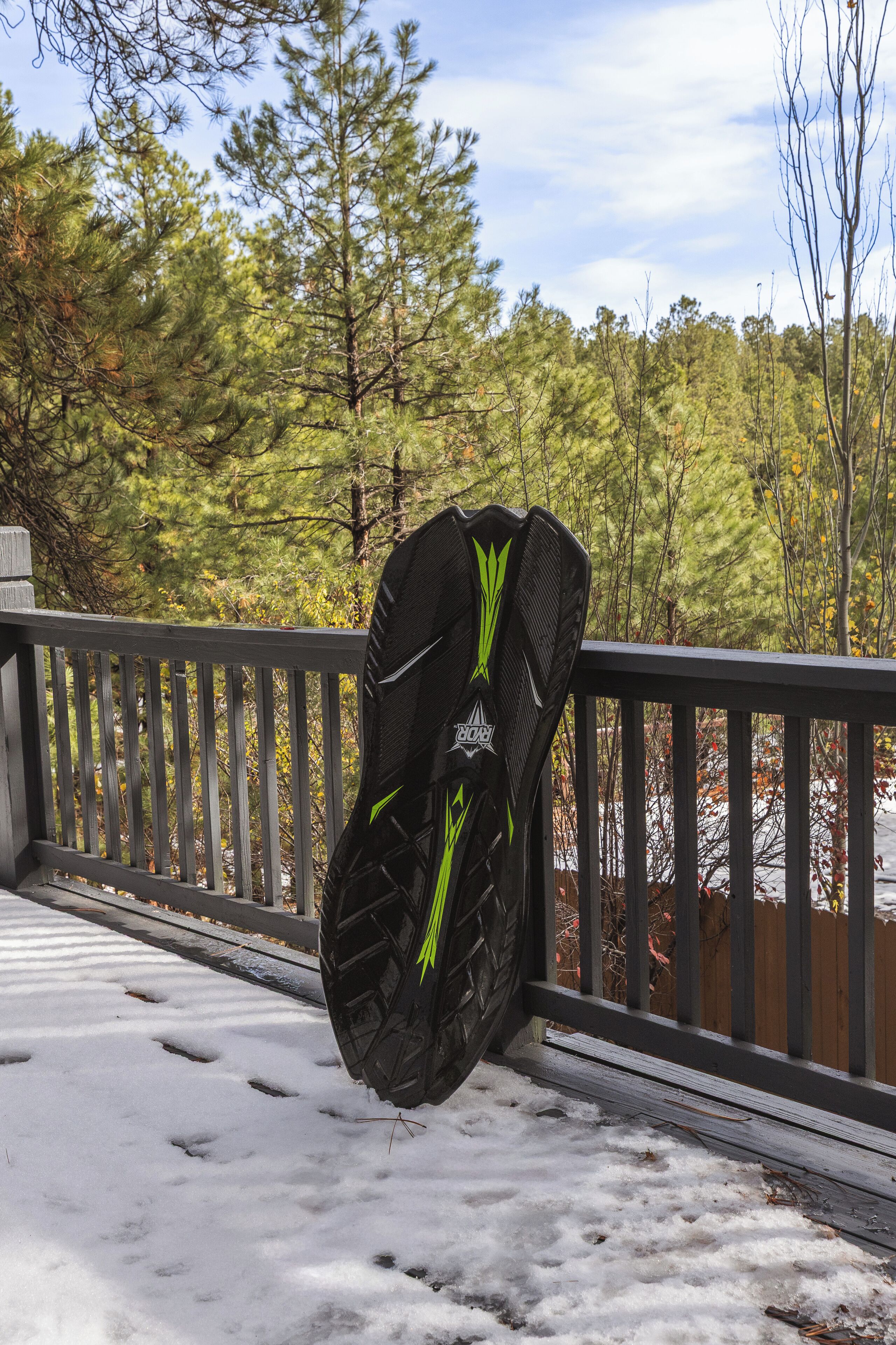 A snowboard leaning against a railing on a snowy deck with a forest in the background.