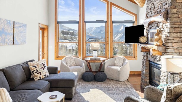 A cozy living room with a stone fireplace, large windows offering mountain views, a flat-screen TV, and comfortable seating. - Black Bear Lodge #303 | Walk to West Wall Lift, Heated Garage + Hot Tub | 2BR (Crested Butte)