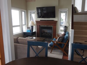 A living room with a fireplace, a television mounted above it, a sofa, and a dining table. - Cosy 2 bedroom with expansive Ocean view and massive deck (Otter Bay)