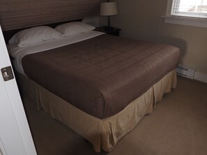 A neatly made bed with a brown bedspread and a beige skirt. There is a nightstand with a lamp and a window with blinds. - Cosy 2 bedroom with expansive Ocean view and massive deck (Otter Bay)