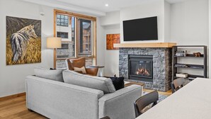 Ein modernes Wohnzimmer mit einem grauen Sofa, einem Holzstuhl, einem Kamin und einem an der Wand befestigten Fernseher.