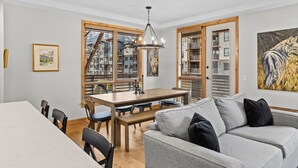 Ein modernes Wohnzimmer mit einem grauen Sofa, einem Holzesstisch und Stühlen. Es gibt ein großes Fenster mit Jalousien, ein Gemälde an der Wand und eine Pendelleuchte.