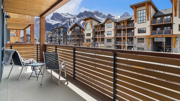Ein Balkon mit Holzgeländer, einem Tisch und zwei Stühlen mit Blick auf ein Gebirge und ein Gebäude mit mehreren Balkonen.