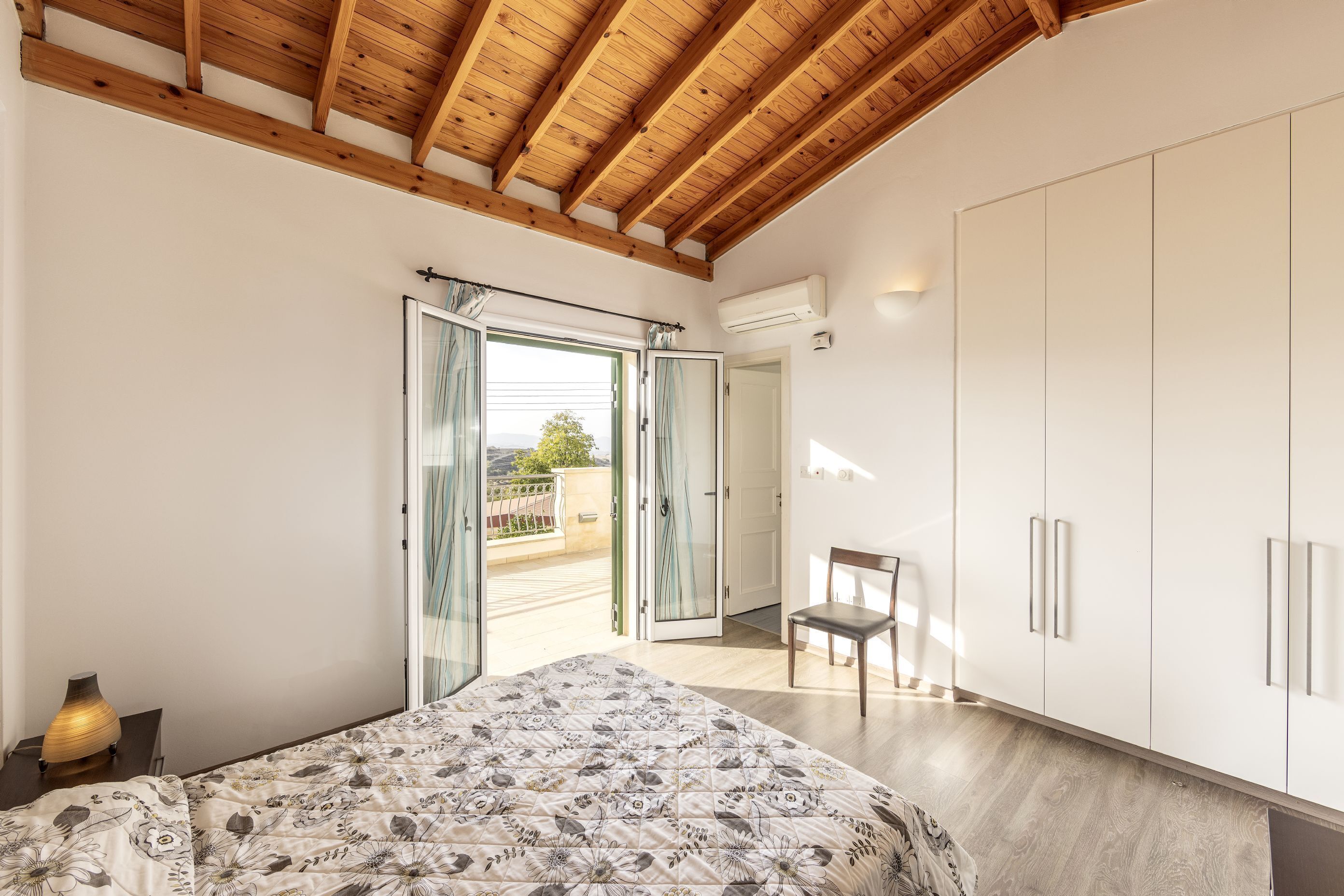 A bedroom with a wooden ceiling, a bed with a floral duvet, a chair, and a wardrobe.