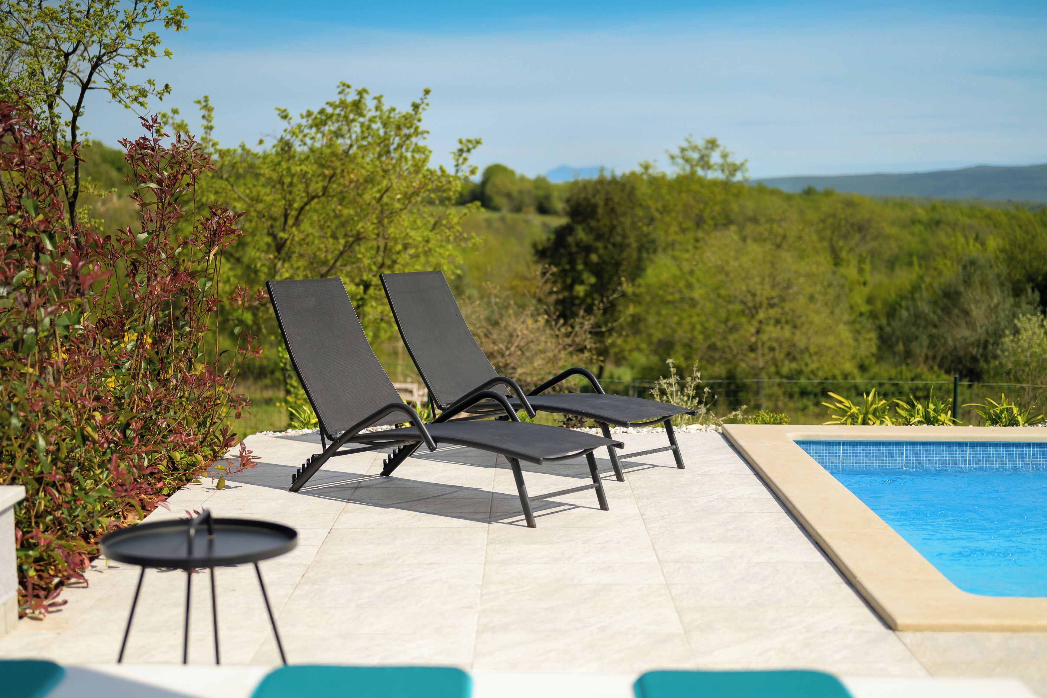 Ein Poolbereich mit zwei Liegestühlen und einem kleinen Tisch, umgeben von einer gefliesten Terrasse und einem Schwimmbecken.