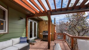 A covered patio with a wooden pergola, a grill, a sofa, and a glass table.