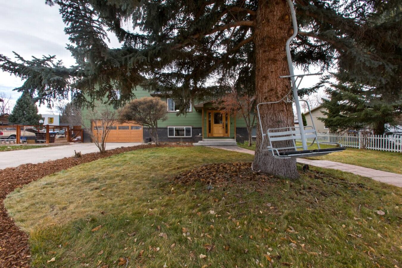 A residential area with a large tree, a bench, and a house with a green exterior and a garage.