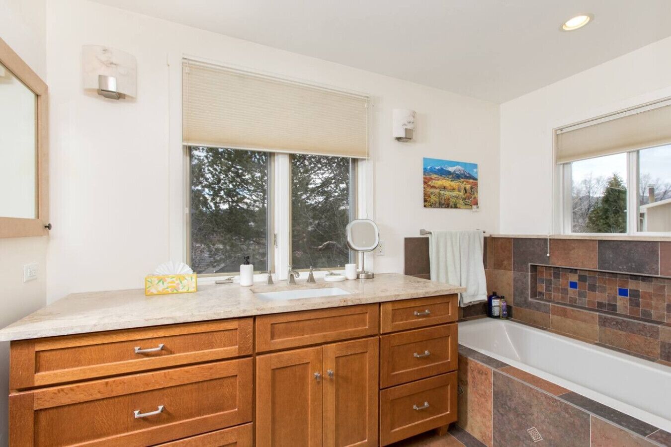 A bathroom with wooden cabinets, a large window, a mirror, and a tiled shower area.