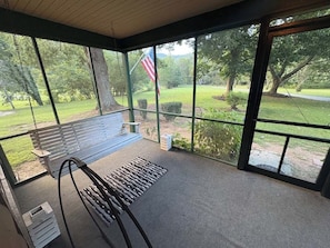 En overdækket veranda med en bænk, et lille bord og udsigt over en græsplæne med træer og et flag.
