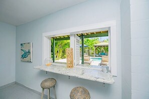 A room with a window overlooking a pool area, a framed artwork on the wall, and two wicker stools. - Decked Out: Steps to Bay & Beach, Pool, Games by Anna Maria Life Vacation Rentals (Anna Maria)