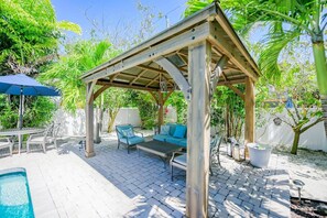 A covered outdoor seating area with a pool, surrounded by lush greenery and a white fence. - Decked Out: Steps to Bay & Beach, Pool, Games by Anna Maria Life Vacation Rentals (Anna Maria)