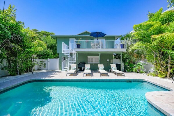 A two-story house with a swimming pool, a balcony with a blue umbrella, and a white fence. - Decked Out: Steps to Bay & Beach, Pool, Games by Anna Maria Life Vacation Rentals (Anna Maria)