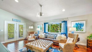 Ein geräumiges Wohnzimmer mit einer großen Couch, zwei Sesseln, einem Couchtisch und einem Deckenventilator. Zwei Glastüren, ein Fenster mit blauen Vorhängen und ein gerahmtes Kunstwerk sind zu sehen.